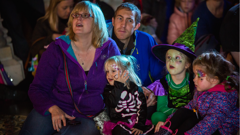 An audience including an enthralled family of five, the children are in Hallowe'en costumes and have painted faces.